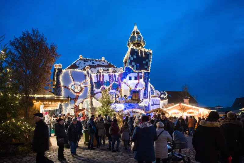 Beleuchtetes Hundertwasser-Kunsthaus in Abensberg mit Weihnachtsmarkt zur blauen Stunde